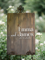A rustic wooden wedding sign on a metal easel stand, featuring the names 'Emma and James' and the date '28.10.