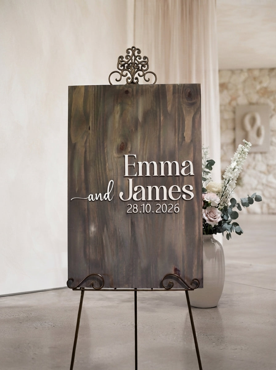 A rustic wooden wedding sign on a metal easel stand, personalized with the names 'Emma and James' and the date '28.10.