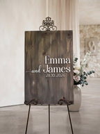 A rustic wooden wedding sign on a metal easel stand, personalized with the names 'Emma and James' and the date '28.10.