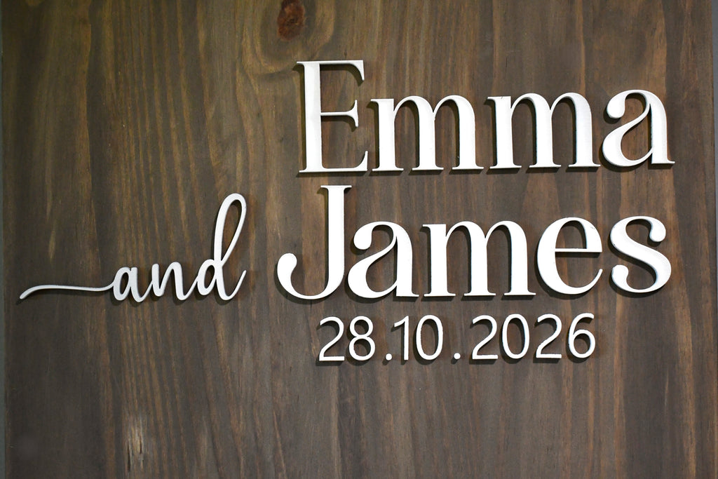 A close-up of a dark brown wooden sign with raised white lettering that reads 'Emma and James 28.10.2026'.