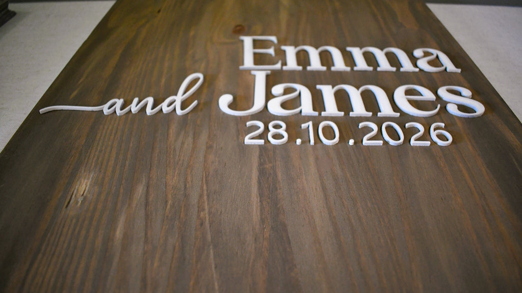 A close-up of a dark brown wooden sign with raised white lettering that reads 'Emma and James 28.10.2026'.